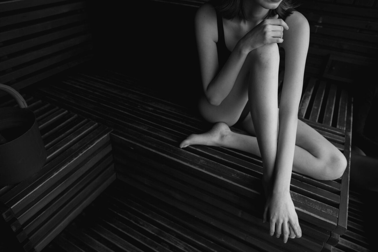A woman in a bathing suit sits elegantly on wooden sauna benches in grayscale.