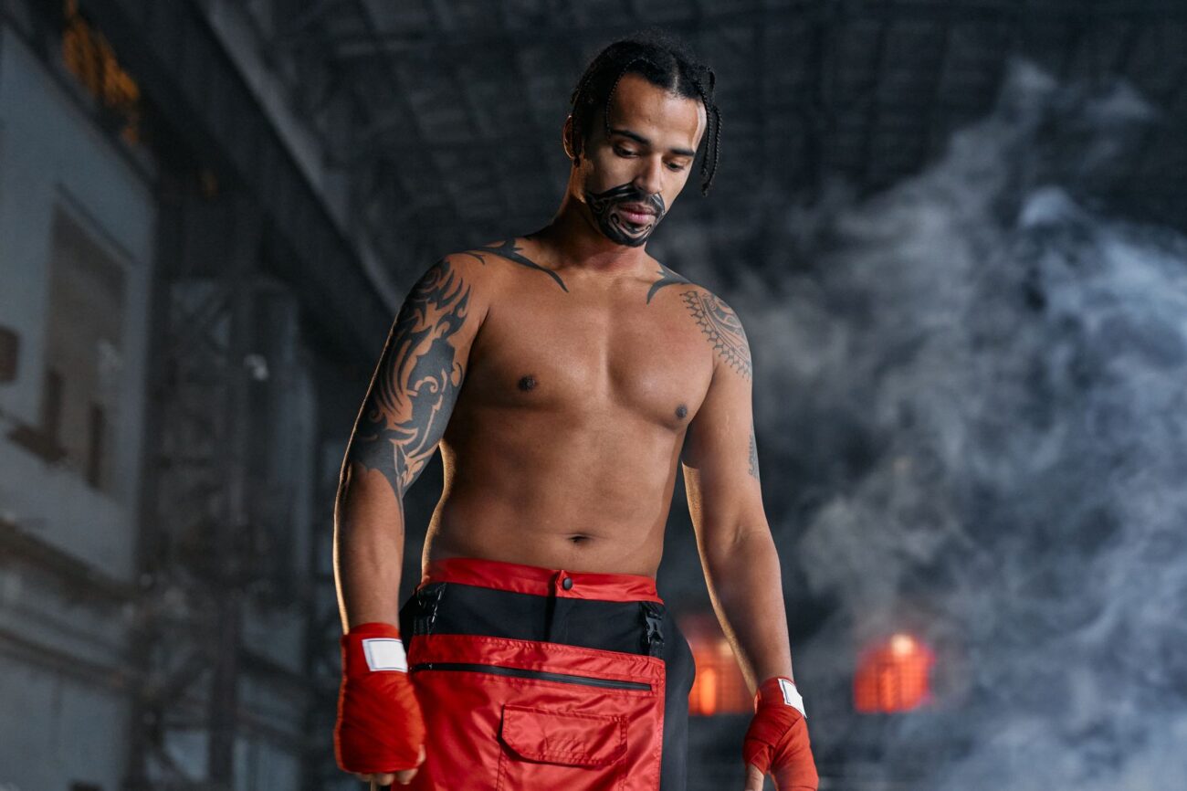 Shirtless tattooed fighter wearing boxing gloves stands amidst smoke indoors.