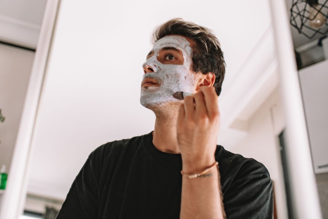 Adult man applying a facial mask while looking in the mirror at home, promoting skincare routine.